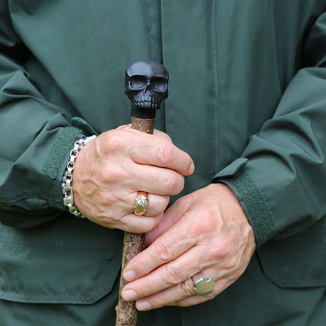 Close-up of hands holding a walking stick with a skull handle, wearing a green jacket.